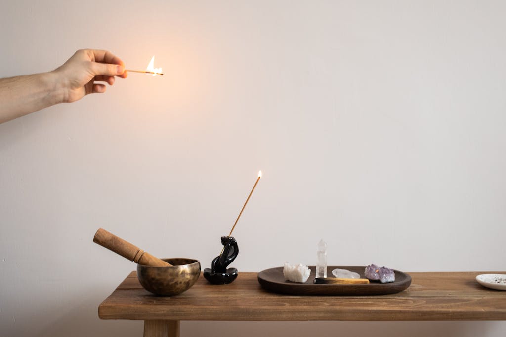 A hand lighting incense near a display of crystals and a mortar on a wooden table.